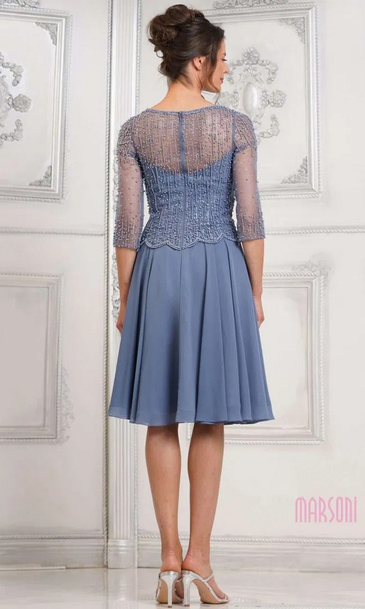 A woman faces away, wearing the Marsoni M312S knee-length semi-formal dress in dusty blue with a sheer beaded bodice and three-quarter sleeves, paired with white heels.